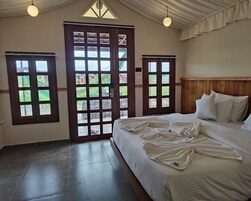 A bedroom with a large bed, wooden headboard, and two windows with views of greenery.