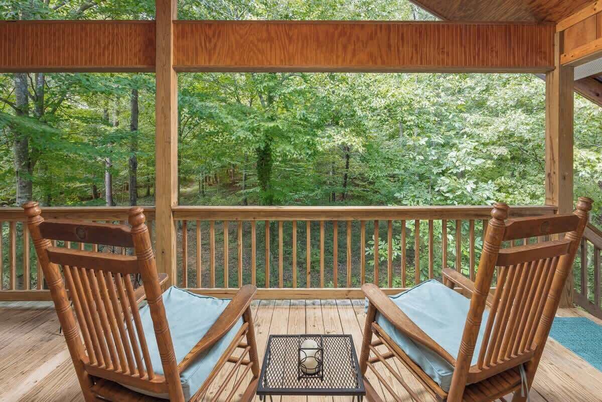 A wooden deck with two wooden chairs and a small table, overlooking a forested area.
