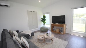 Uma sala de estar moderna com um sofá cinzento, um puff redondo, uma televisão sobre um suporte de madeira e uma grande janela com vista para o exterior.