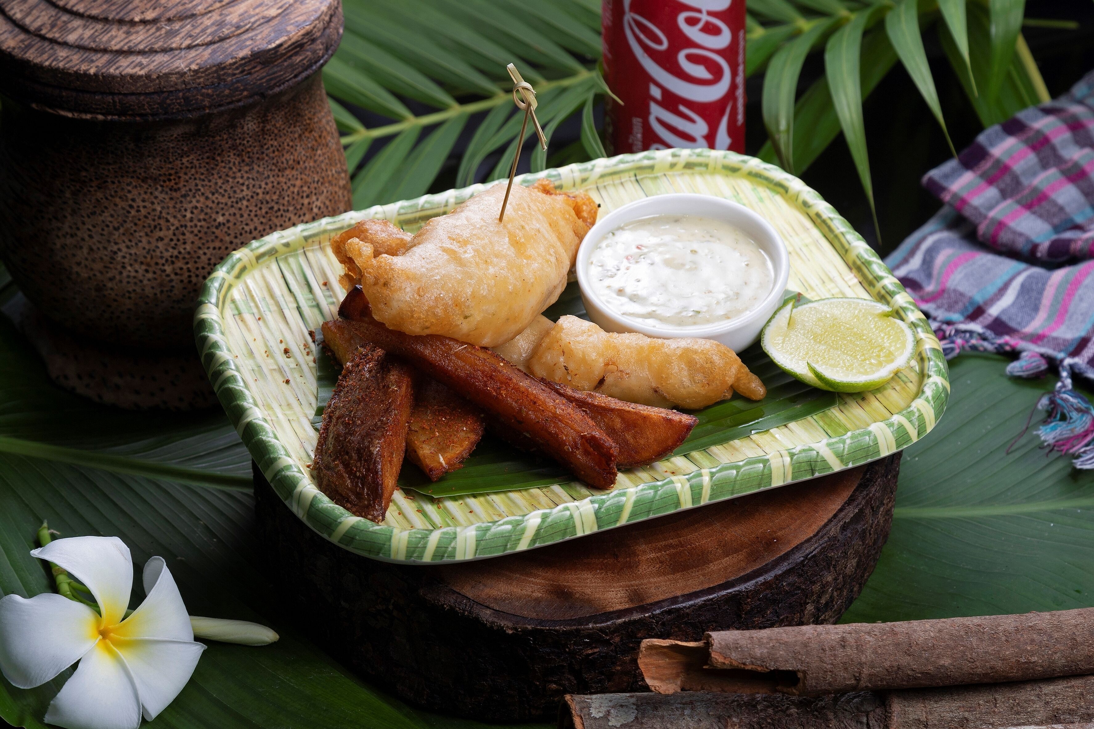 A plate of fried fish and chips with a side of dipping sauce and lime, set on a wooden table with a banana leaf.