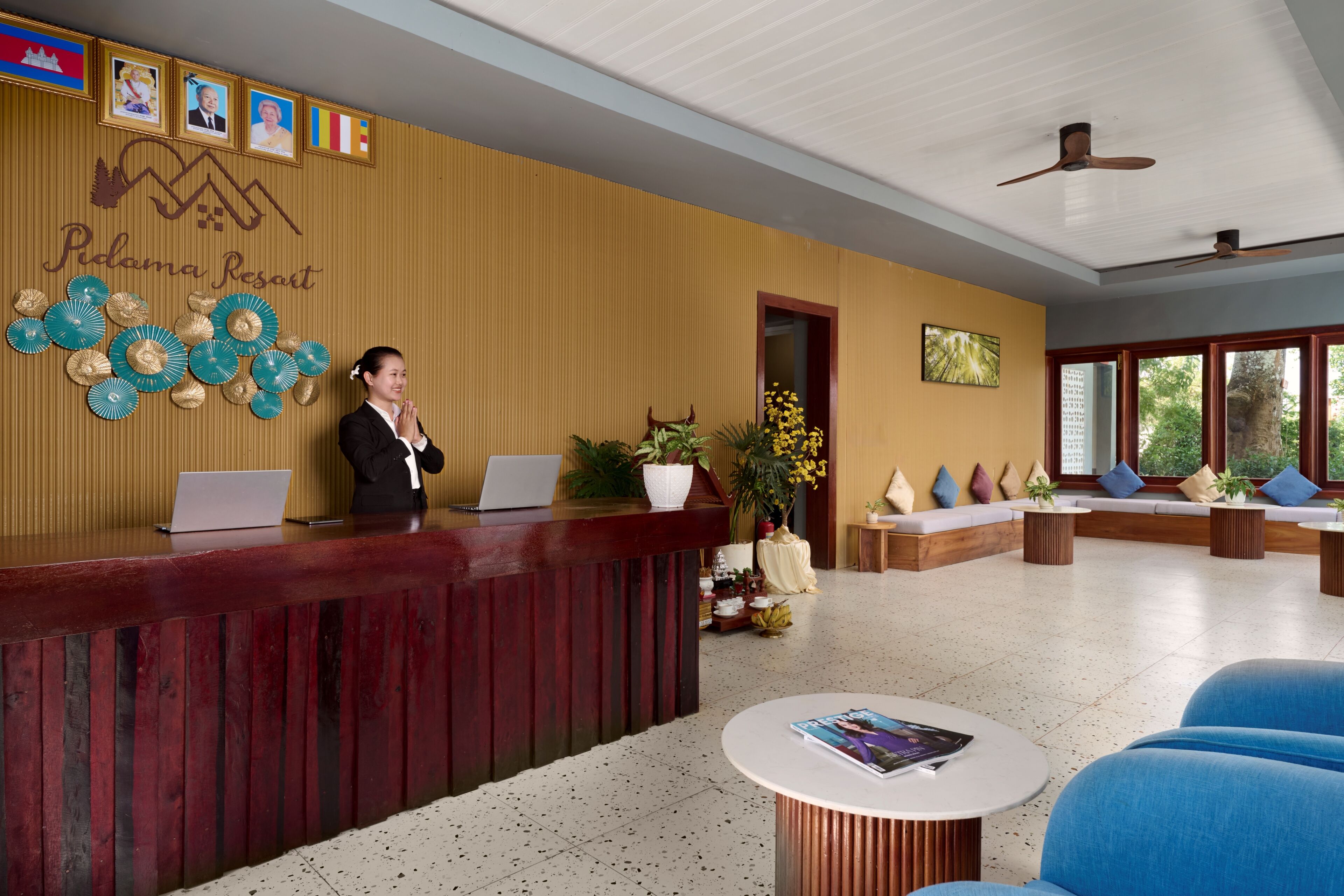 A reception desk with a laptop, a woman standing behind it, and a seating area with sofas and a coffee table.