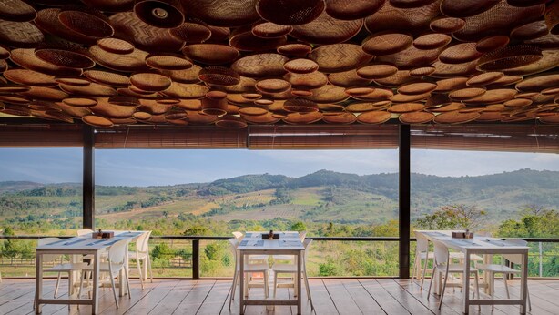 Una terraza con piso de madera, mesas y sillas blancas, y vistas a colinas verdes.
