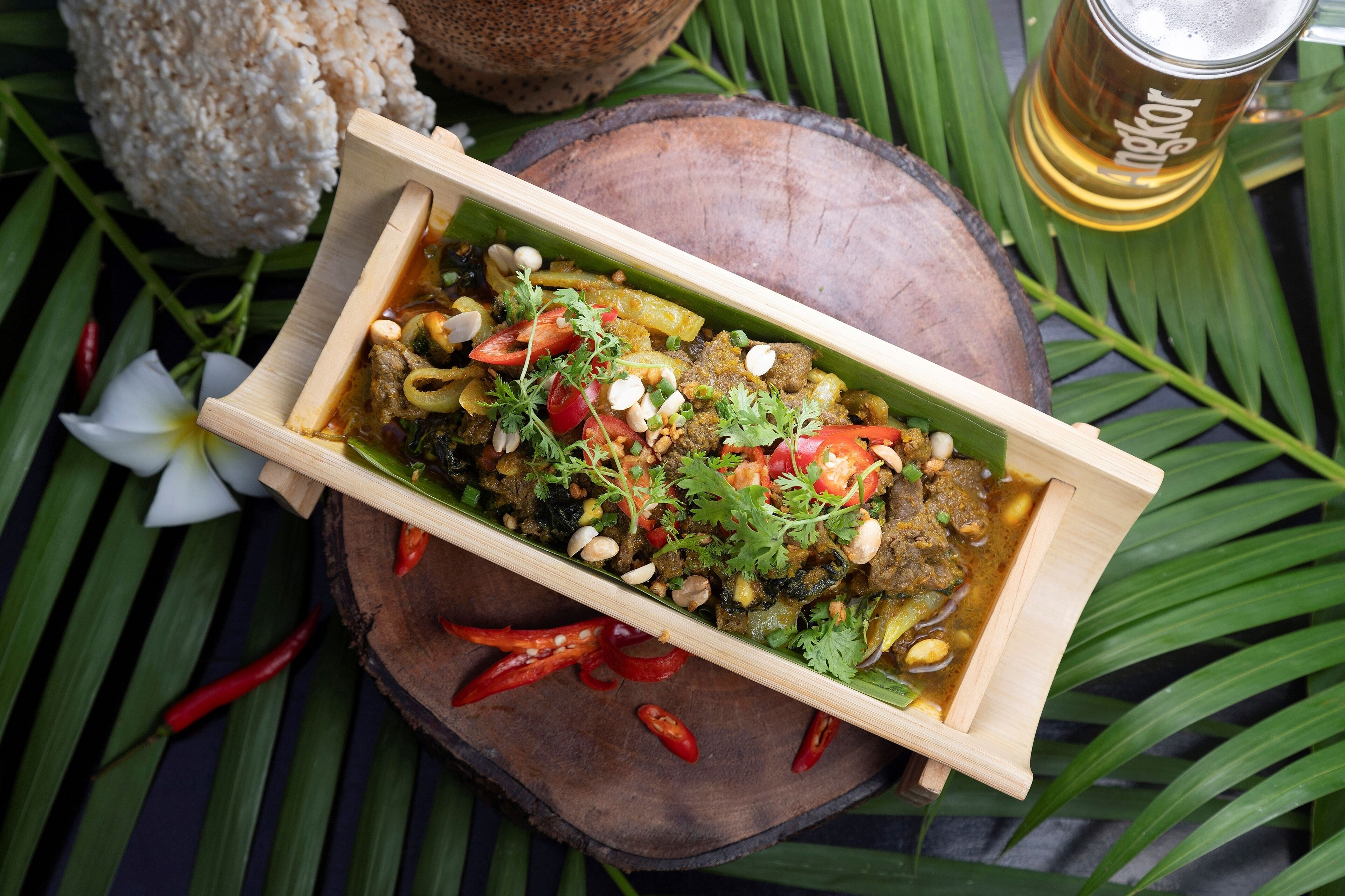 Un plat de service en bois contenant un plat de curry, entouré de feuilles vertes et de piments rouges.