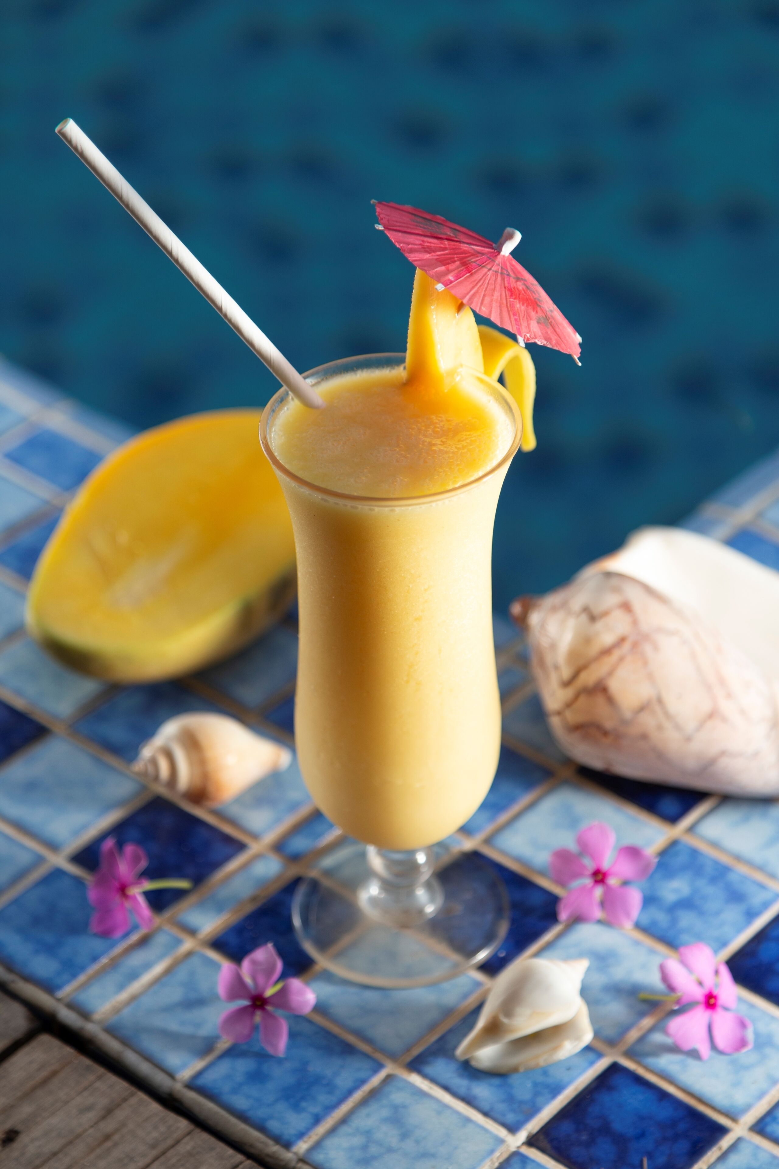 A glass of tropical drink with a straw and a red umbrella garnish, a half mango, and a coconut on a blue tiled surface.