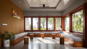 Una habitación amplia con ventanas grandes, un ventilador de techo y un área de descanso con mesas redondas de madera.