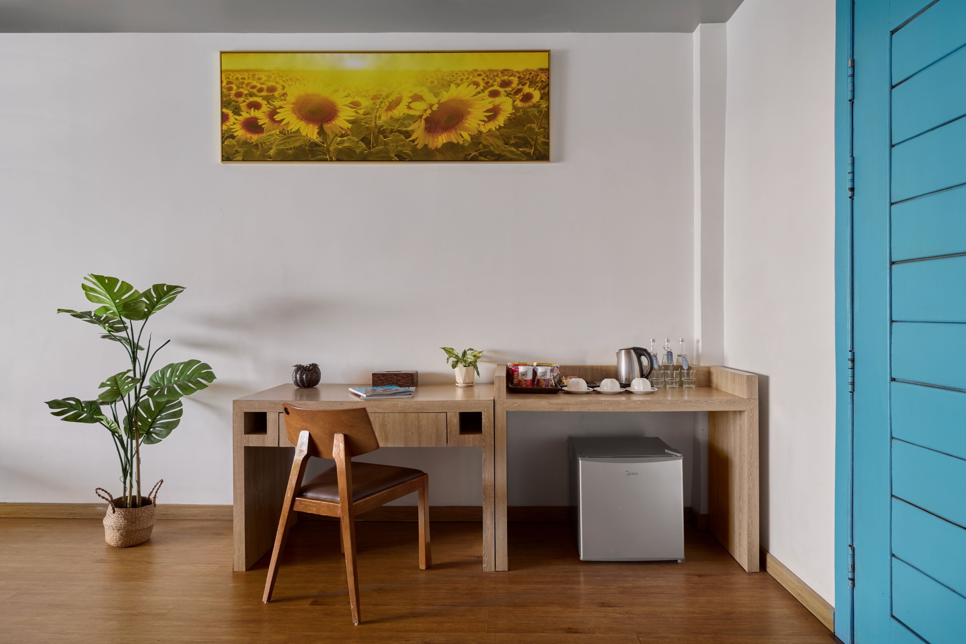 Een kamer met een houten bureau, een stoel, een kleine koelkast, een potplant en een schilderij van zonnebloemen aan de muur.