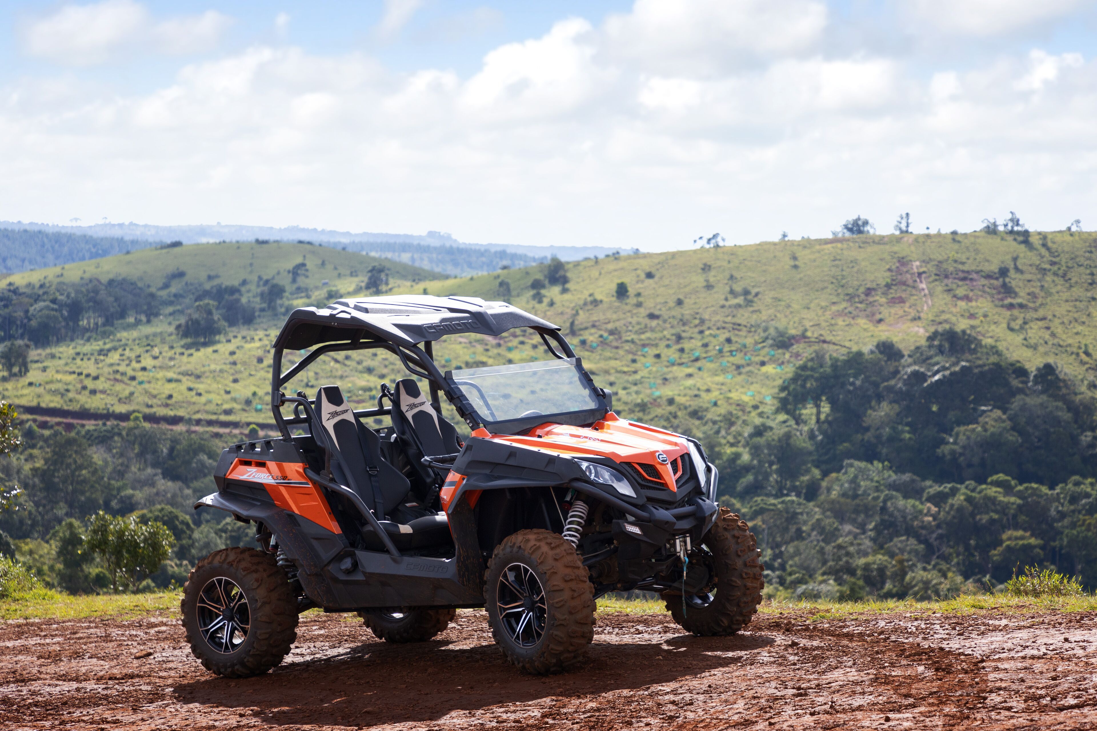A red and black off-road vehicle parked on a dirt path with a scenic view of green hills and trees in the background.