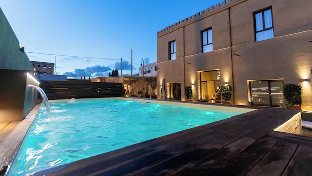 Una piscina con muelle de madera, un edificio moderno con grandes ventanales y un cielo despejado.