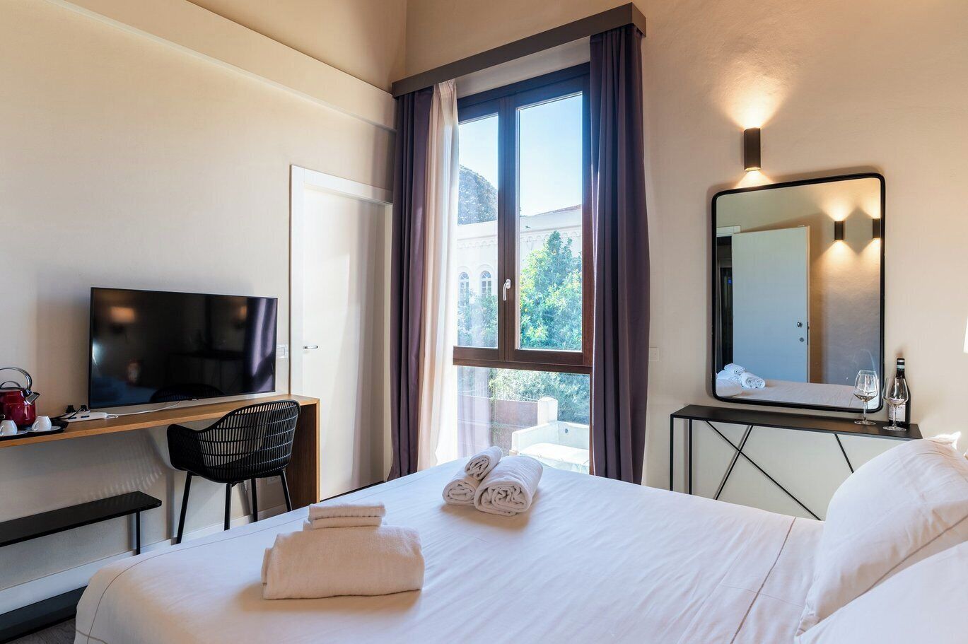 Quarto de hotel com cama, escrivaninha com televisão, poltrona, janela com cortinas e espelho.