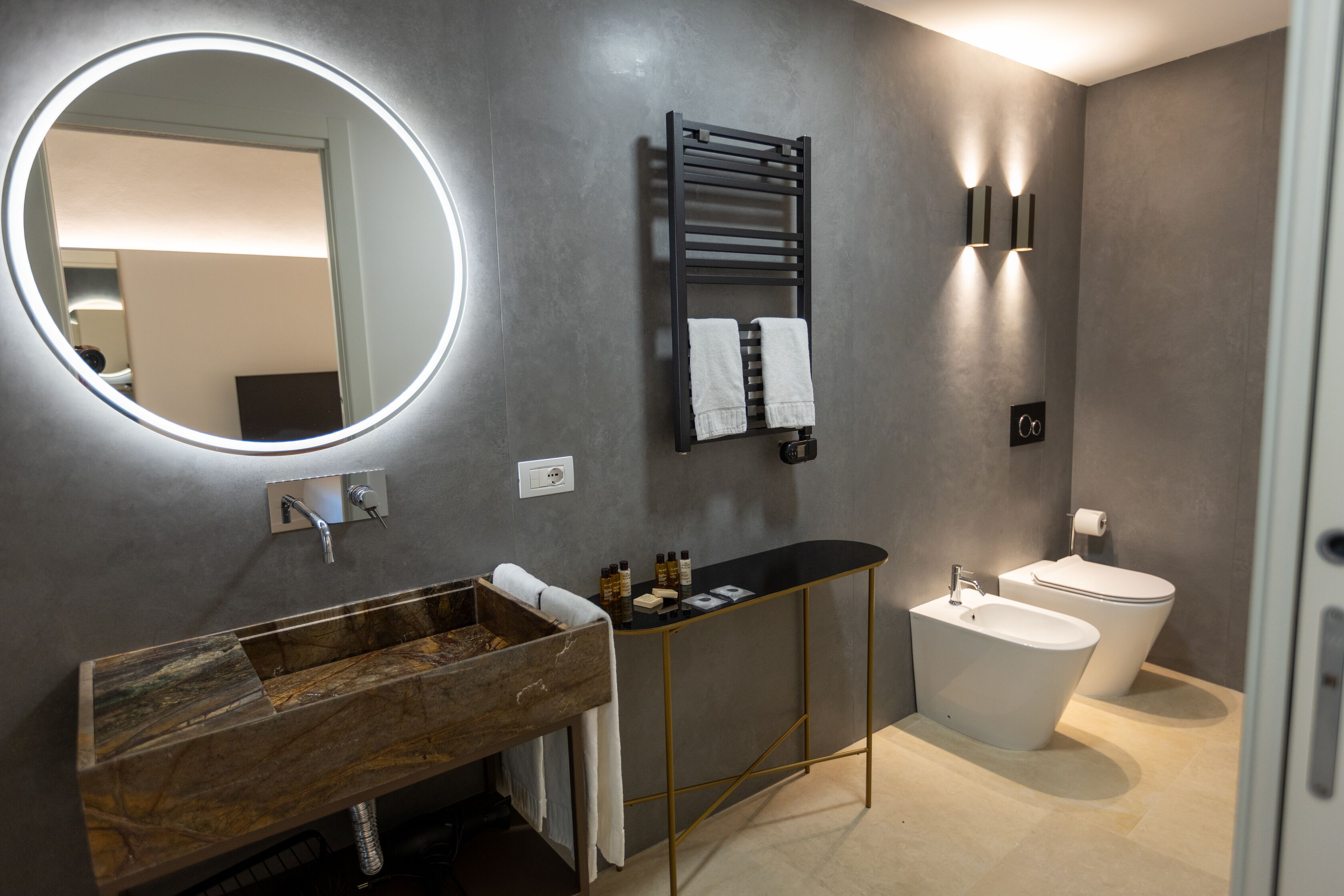 A modern bathroom with a wooden vanity, a round mirror, a towel rack, a toilet, and a bidet.