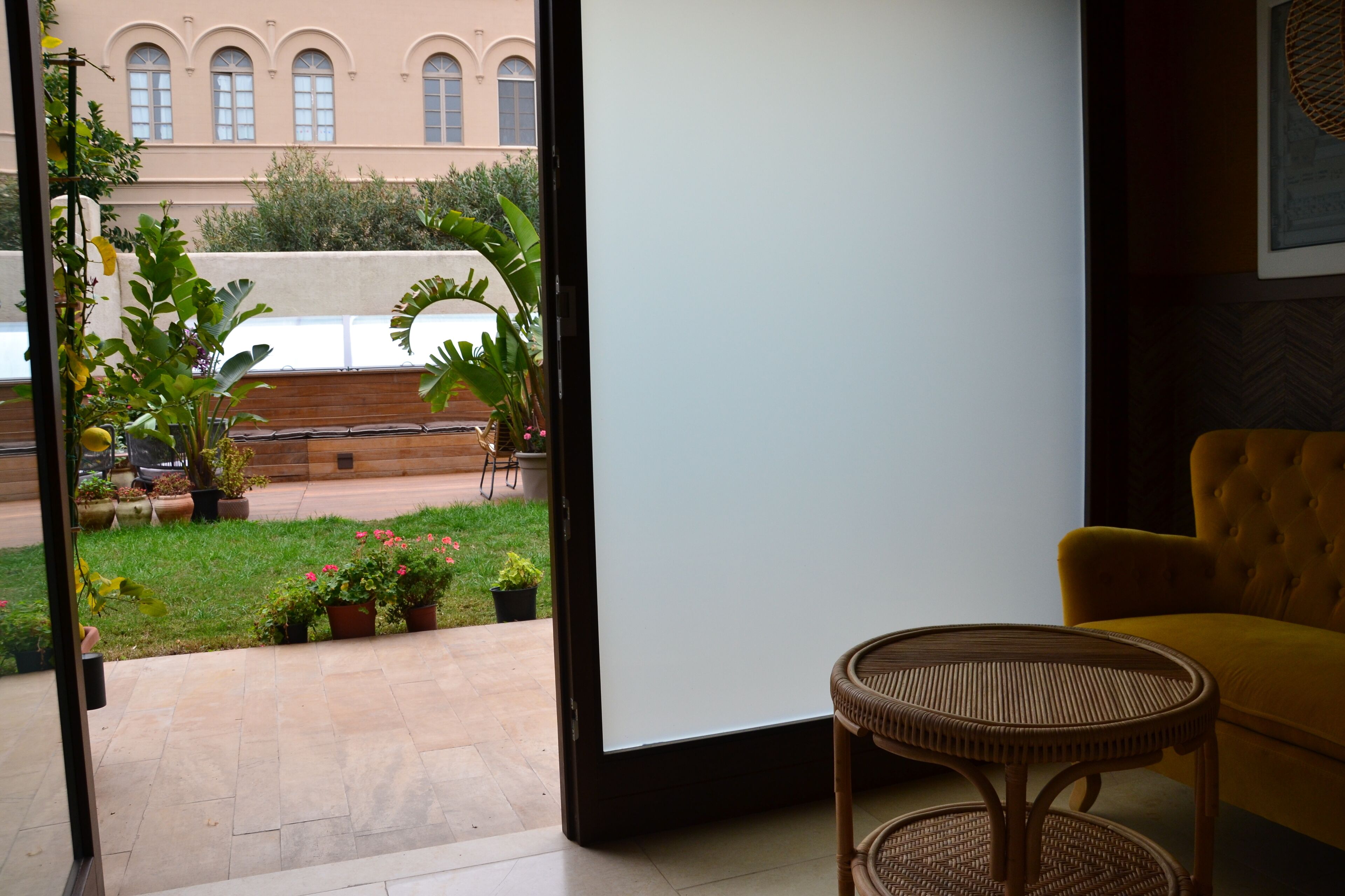 A room with a view of a garden and a building. There is a wicker chair and a yellow sofa.