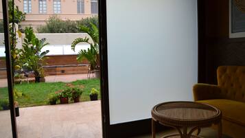 A room with a view of a garden and a building. There is a wicker chair and a yellow sofa.