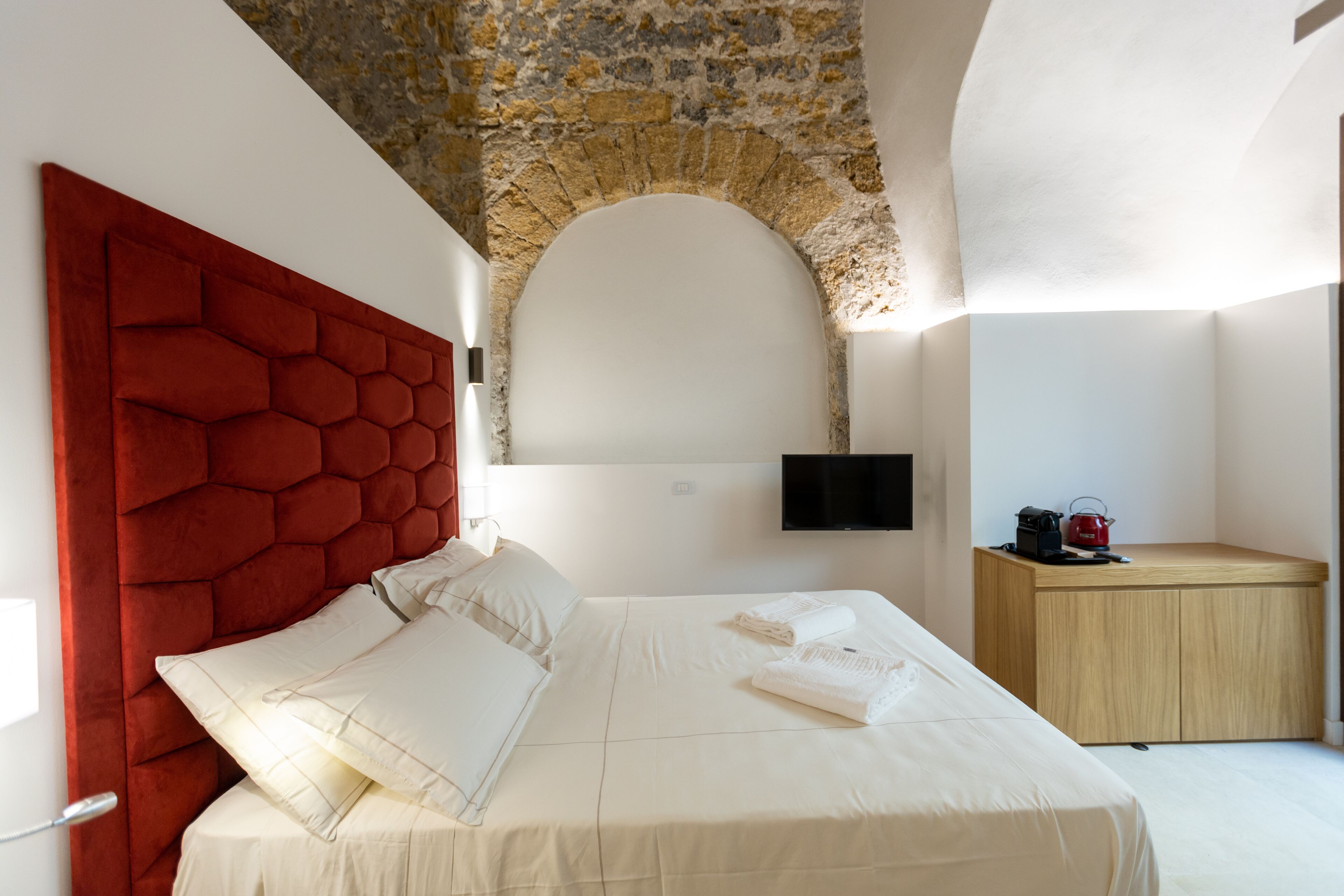 A modern bedroom with a stone arch ceiling, a red upholstered headboard, a wooden bedside table with a kettle, and a flat-screen TV mounted on the wall.