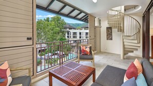 Ein Balkon mit Blick auf einen Pool und Gebäude, ein Holztisch, Korbstühle und eine Wendeltreppe.