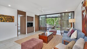 Ein geräumiger Wohnbereich mit einer Couch, Hocker und einem Couchtisch. An der Wand befindet sich ein Fernseher und ein Gemälde.