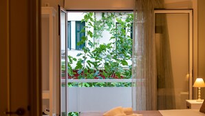 A hotel room with a bed, a window with a view of greenery, a bedside table with a lamp, and a door leading to another room.