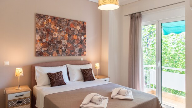 A hotel room with a large bed, two bedside tables with lamps, a decorative wall art piece, and a balcony with a view of greenery.