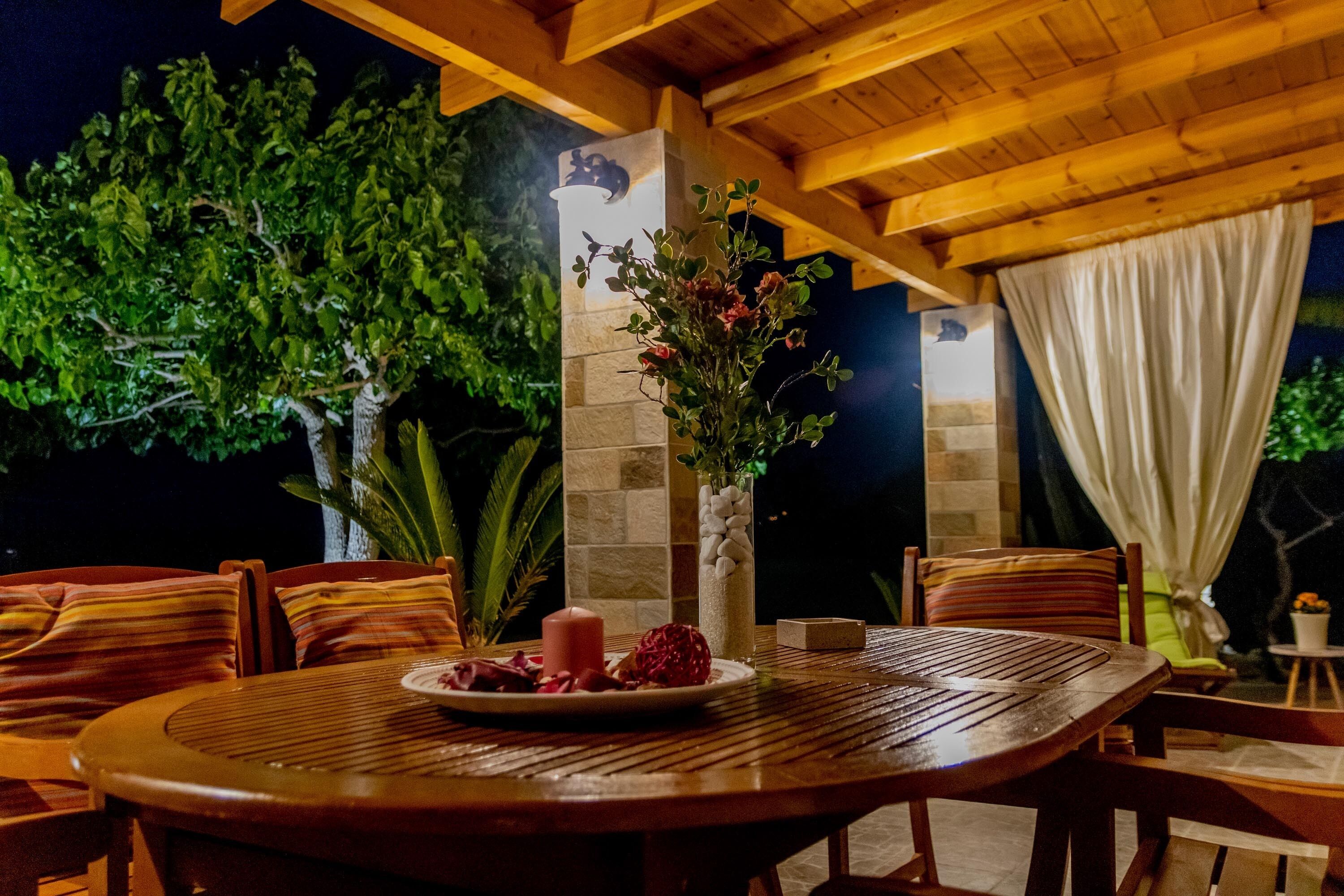 An outdoor dining area with a round wooden table, chairs, a vase with flowers, and a stone pillar with a light fixture.
