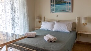 Una cama bien hecha con almohadas blancas y una colcha azul, un cuadro de una escena costera, un aire acondicionado montado en la pared y una mesita de noche con una lámpara.
