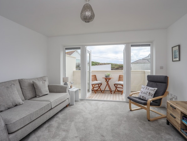 A modern living room with a grey sofa, a wooden chair, a small table with a lamp, and a view of the outside balcony.