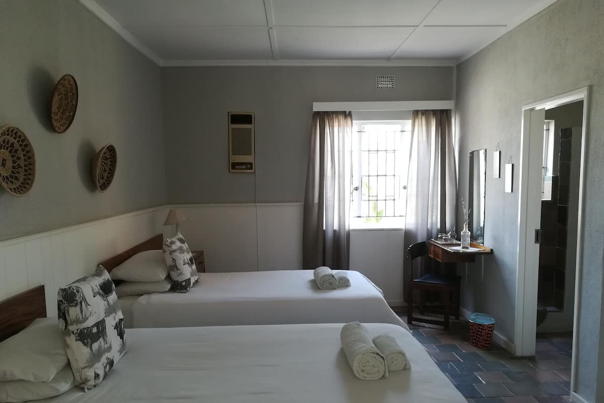 Une chambre d’hôtel avec deux lits, un bureau, une chaise, une fenêtre avec des rideaux et une petite poubelle.