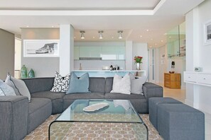 A modern living room with a glass coffee table, grey sofas, and a kitchen in the background. - Horizon Bay 1602 Penthouse Apt with lovely views (Cape Town)