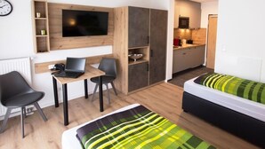 Habitación pequeña con cama, escritorio con computadora portátil, silla, televisor montado en la pared y zona de kitchenette.