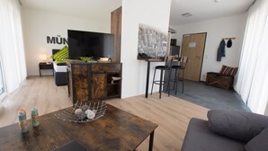 Una sala de estar moderna con sofá, una mesa de centro y televisor. Hay una zona de cocina con mesa y sillas.