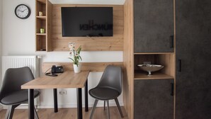 Una habitación moderna con una mesa de comedor de madera, dos sillas negras, un televisor montado en la pared, un pequeño estante con adornos y un armario gris.