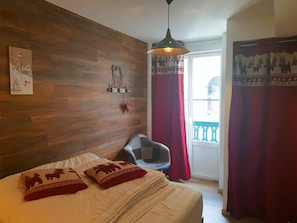 Quarto com cabeceira de madeira, cama com roupa de cama branca e almofadas vermelhas, poltrona cinza, luminária pendente e janela com cortinas vermelhas.