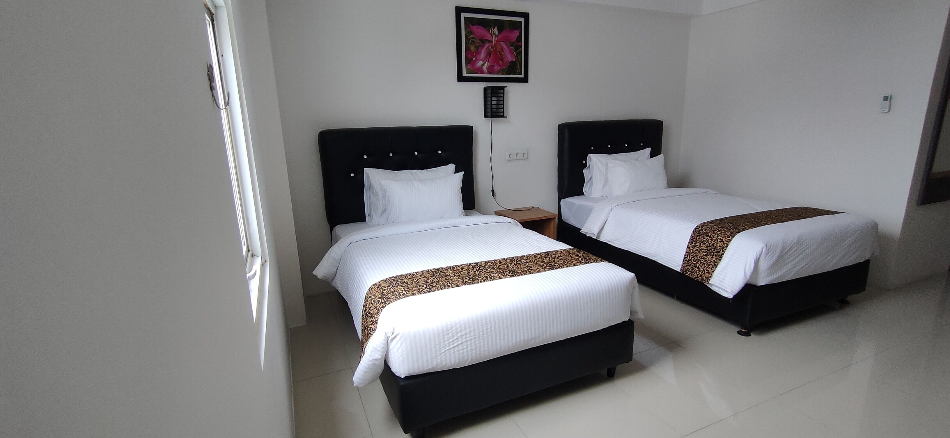 A hotel room with two beds, each with a black headboard and white bedding. There is a small table between the beds, and a framed picture on the wall above the beds.