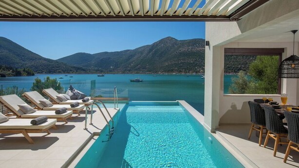 Zona de piscina con sillones de descanso y una mesa, con vista a un lago con barcos y montañas.