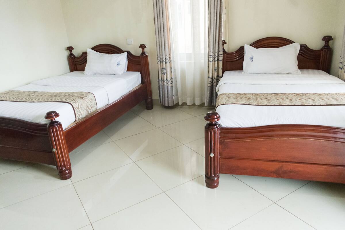 Two wooden beds with white linens and pillows, a tiled floor, and a window with sheer curtains.