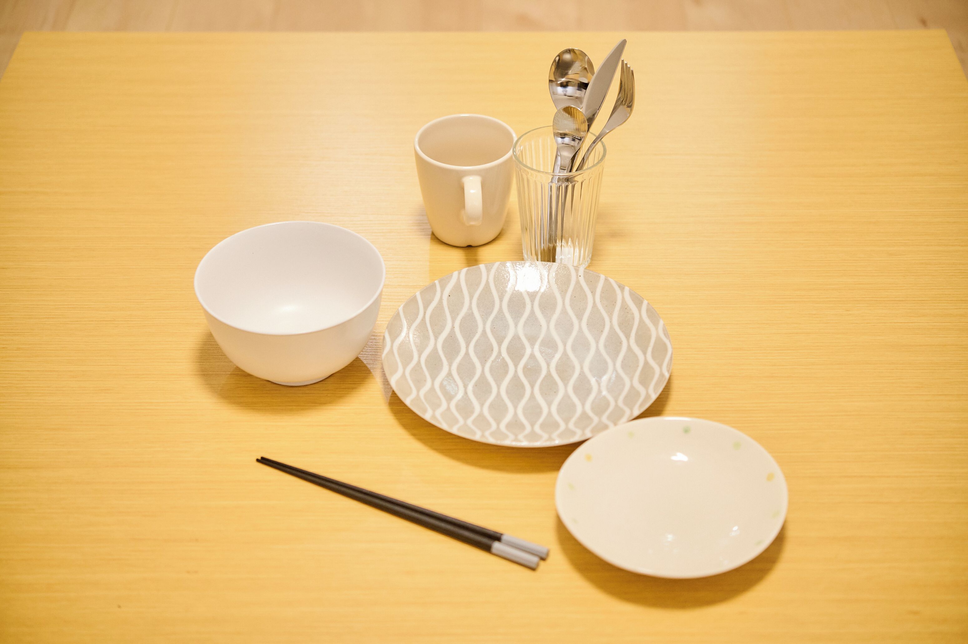A wooden table with a white bowl, a patterned plate, a cup, and chopsticks.