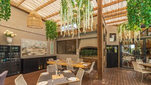 Un restaurant avec des tables en bois et des chaises en osier, des plantes suspendues et un grand tableau au mur.