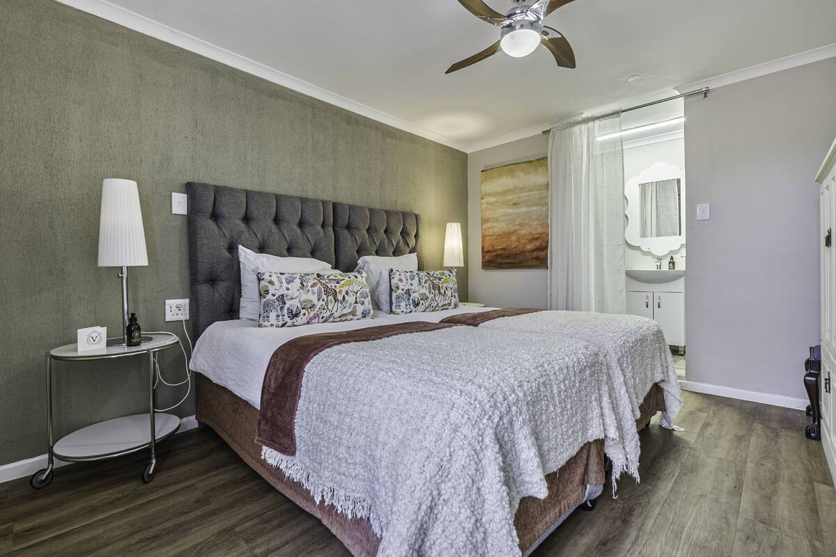 Een slaapkamer met een gestoffeerd hoofdeinde, een bed met gekleurde kussens, een nachtkastje met een lamp en een plafondventilator.