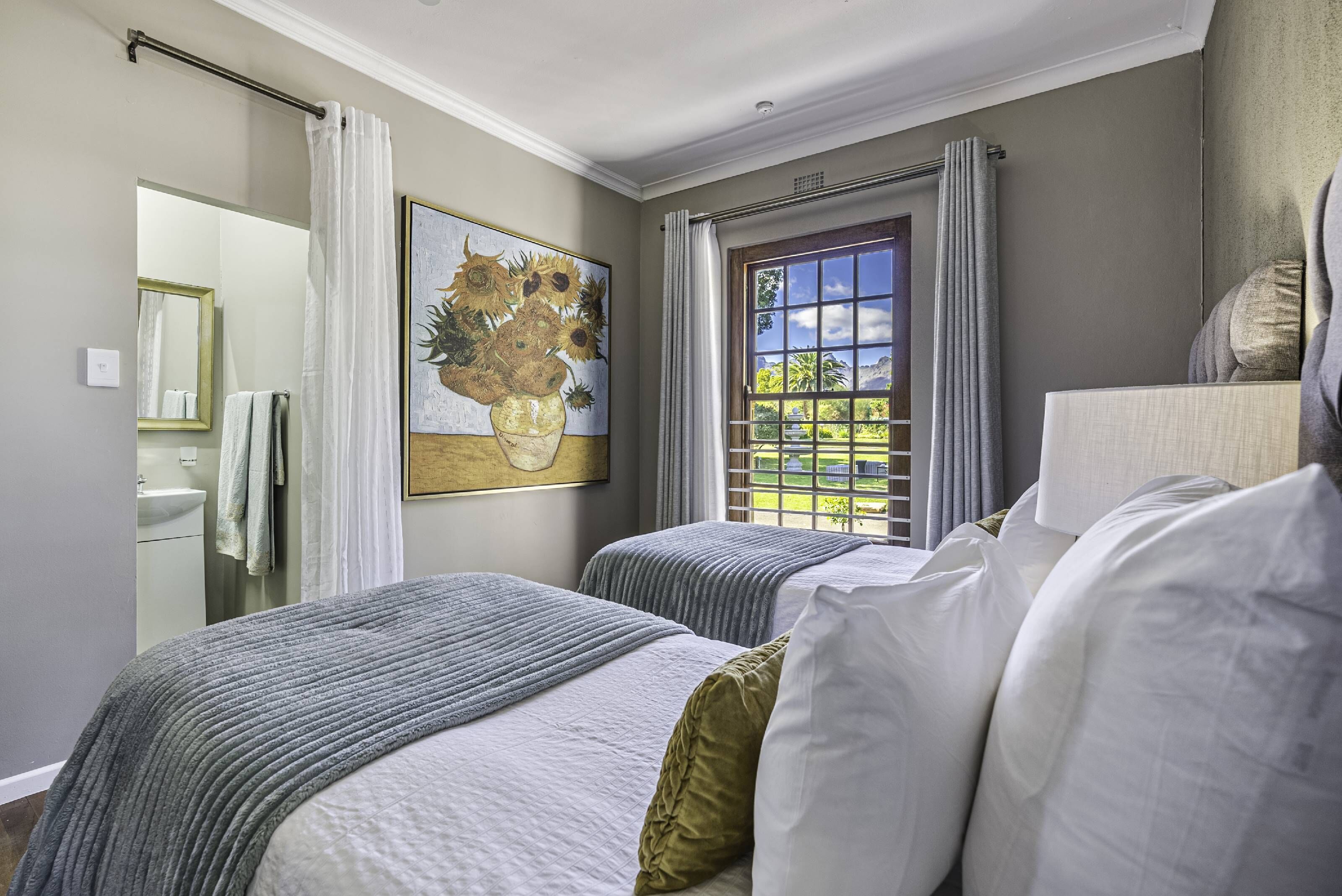 A hotel room with two beds, a large window with curtains, a painting of sunflowers, and a bathroom visible through an open door.