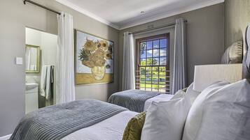 A hotel room with two beds, a large window with curtains, a painting of sunflowers, and a bathroom visible through an open door.