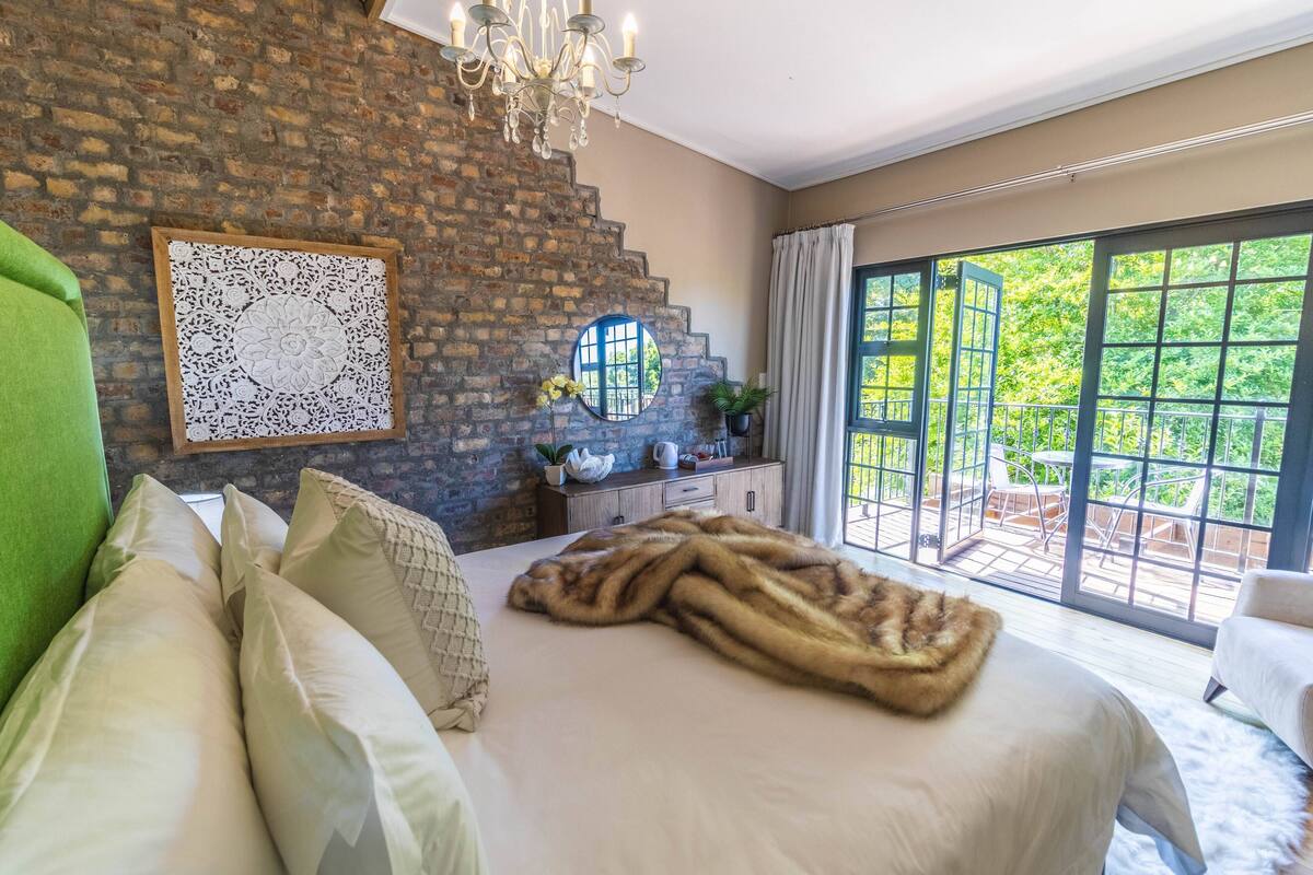 Een slaapkamer met een groot bed, een groene fauteuil, een kroonluchter, een decoratief kunstwerk aan de muur en uitzicht op de buitenwereld door grote ramen.