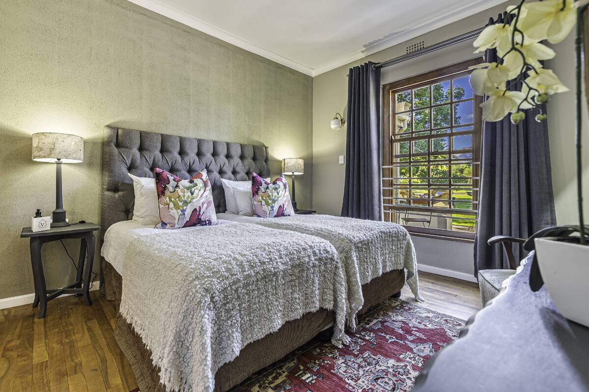 Een slaapkamer met een groot bed, twee nachtkastjes met lampen, een raam met gordijnen en uitzicht op groen buiten.
