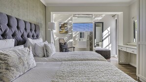 Une chambre à coucher avec une tête de lit capitonnée, un grand lit avec de nombreux oreillers, une table de chevet et une chaise. On y trouve une fenêtre avec des stores, une porte donnant sur une autre pièce et une vue sur un balcon.