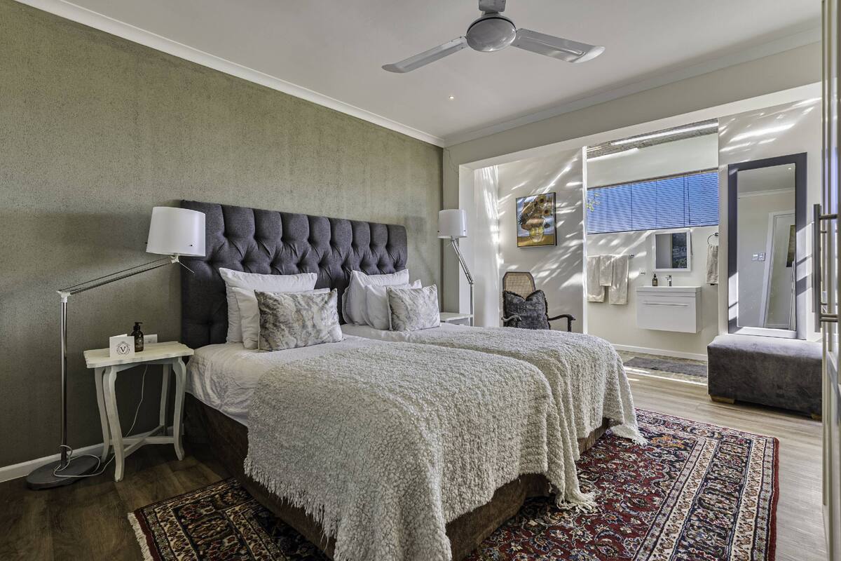 Een slaapkamer met een gestoffeerd hoofdeinde, een groot bed met een witte deken, een bedlampje en een kamerkleed met een patroon.