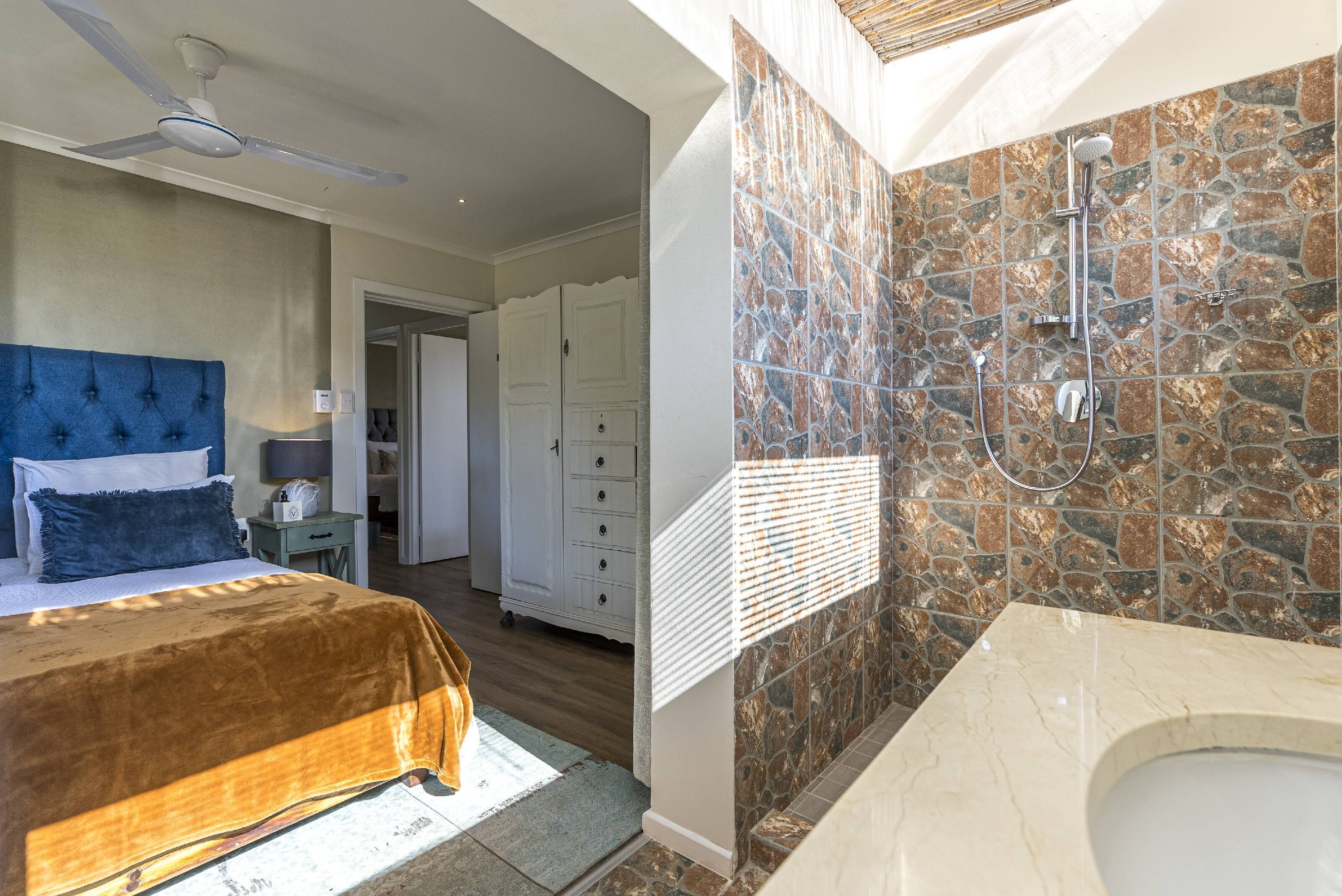 Une chambre dotée d’un grand lit, d’un ventilateur de plafond et d’une salle de bain avec un mur de douche en pierre et une baignoire.
