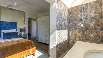 Une chambre dotée d’un grand lit, d’un ventilateur de plafond et d’une salle de bain avec un mur de douche en pierre et une baignoire.