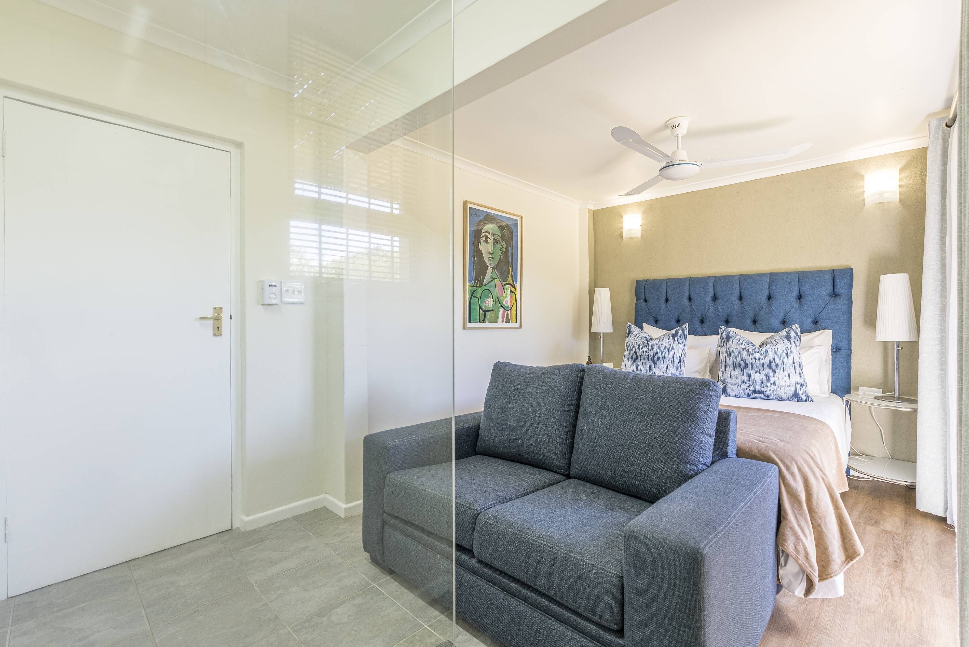 A bedroom with a grey sofa, a bed with a blue headboard, and a ceiling fan.