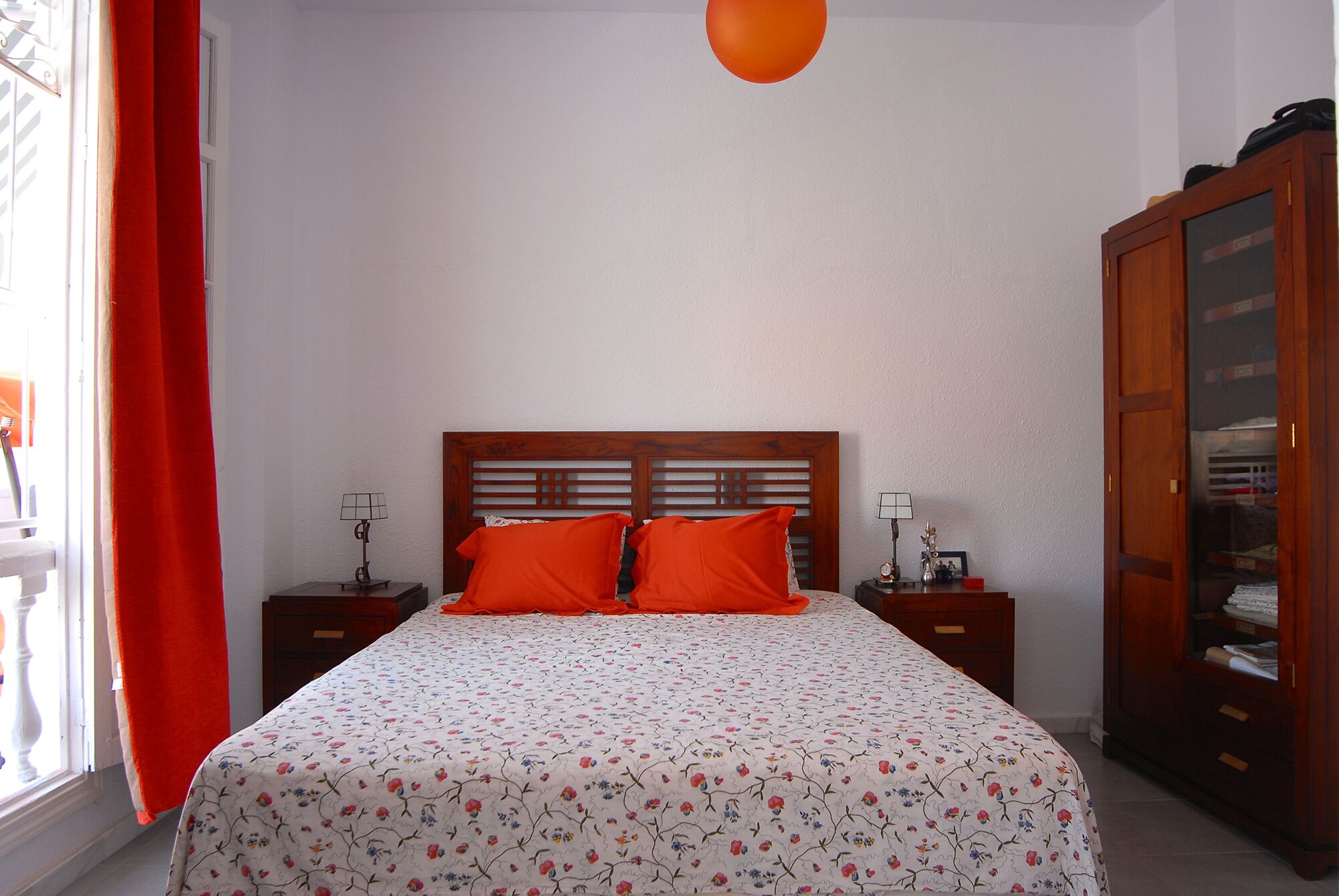 Een slaapkamer met een bed, twee nachtkastjes, een houten garderobe en een raam met een rood gordijn.