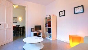 Una sala de estar moderna con una mesa redonda blanca, una estantería, un televisor y una puerta que da a otra habitación.