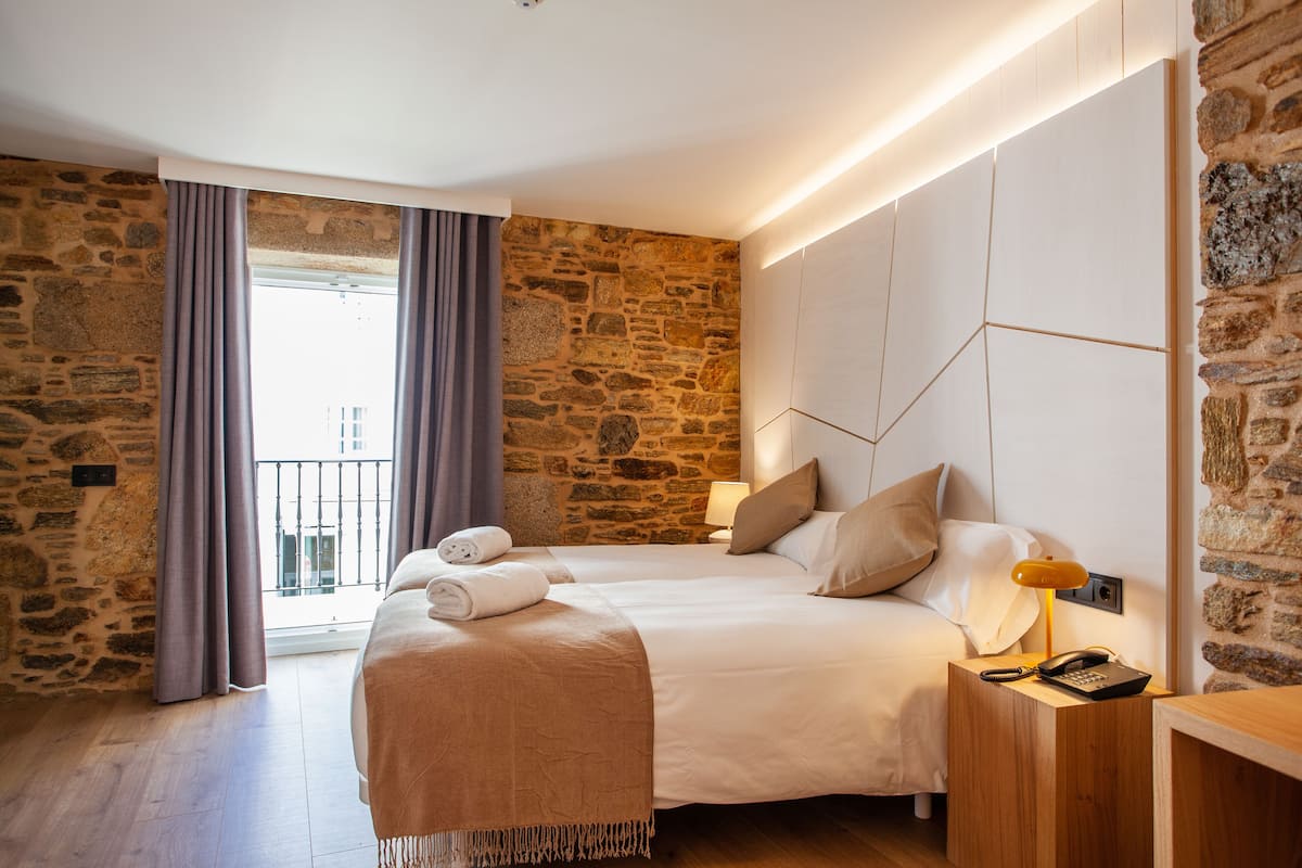 A hotel room with a stone wall, a bed with a wooden headboard, a bedside table with a lamp, and a view of the outside balcony.