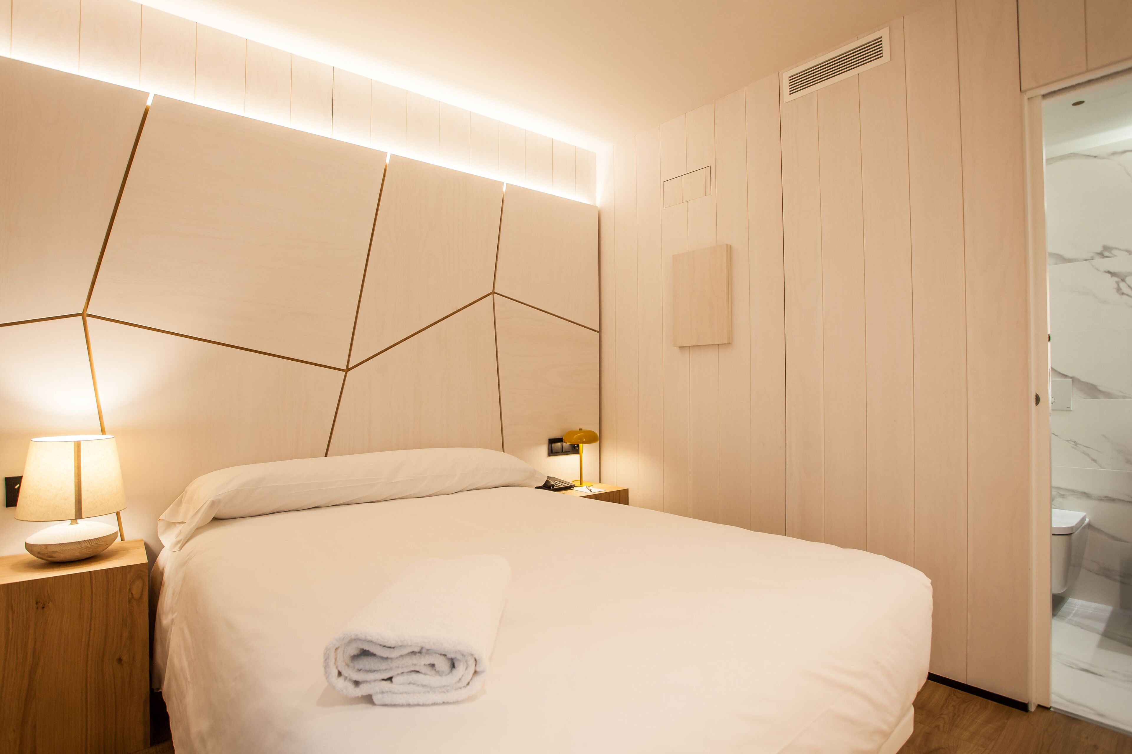 A modern hotel room with a geometric headboard, a neatly made bed with a rolled towel, a bedside lamp, and a visible bathroom with a marble shower.