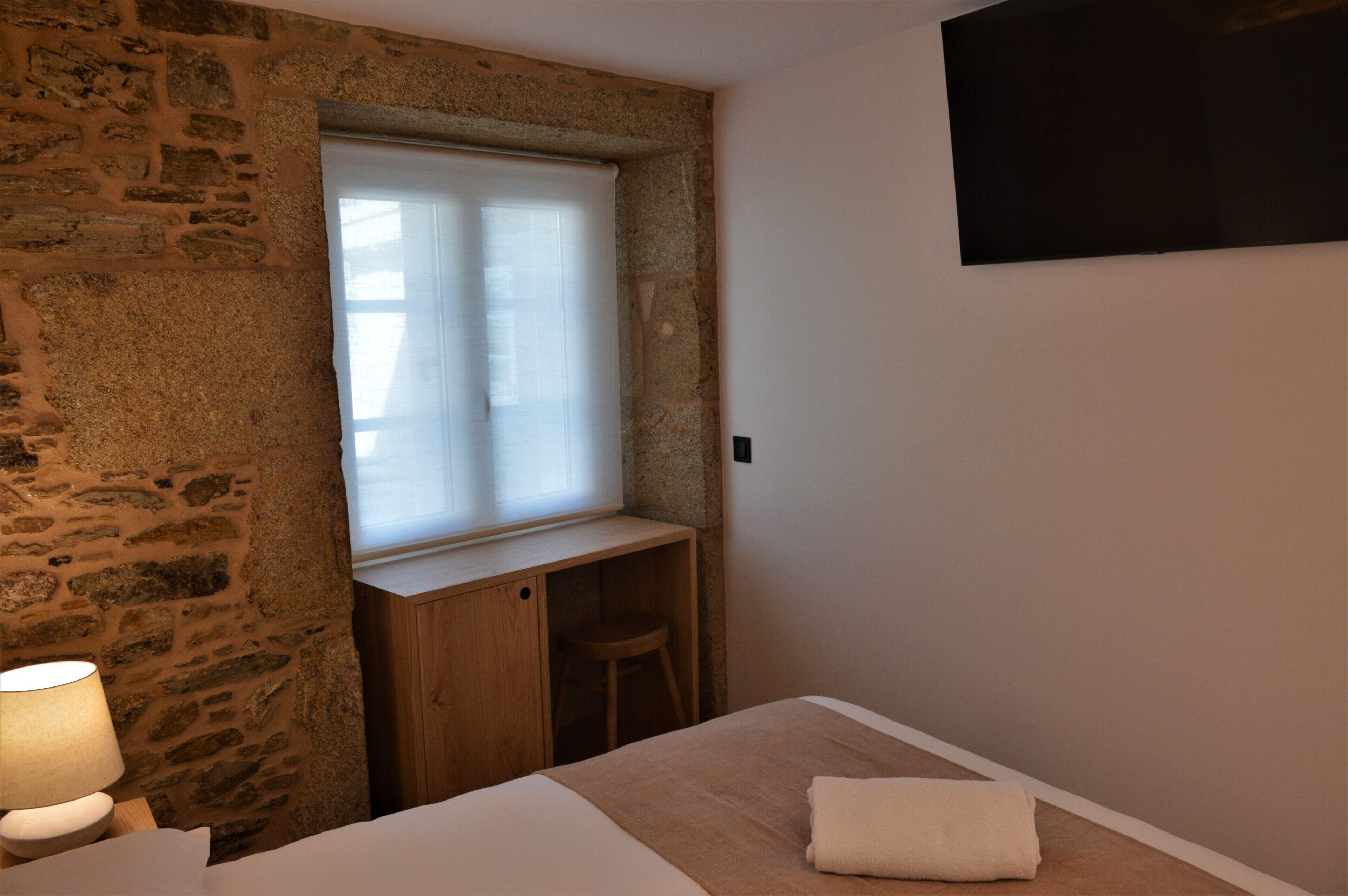 Un dormitorio con pared de piedra, un escritorio de madera, una ventana con persianas, una cama con manta blanca y un televisor fijado a la pared.